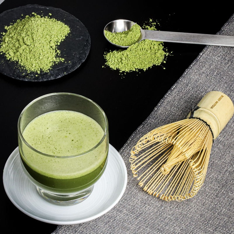 Perfect Glass cup of prepared Matcha drink (latte) and the Chasen (bamboo whisk) for purpose of matcha tea  laying on side of it with black and grey background. Pure finely ground spoon of Matcha green tea powder.  ‘Enjoy Various benefits’ with plant based recipe and good quality traditional style whisk.